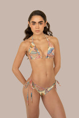 A woman modeling a two-piece string bikini with a colorful tropical leaf print. The bikini features a triangle top with a central ring detail and matching side-tie bottoms, set against a plain beige background.