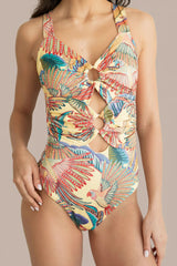 A woman wearing a one-piece swimsuit with a tropical leaf and bird print on a cream background. The swimsuit features a halter-style neckline with a keyhole cutout at the chest and a side cutout near the waist.