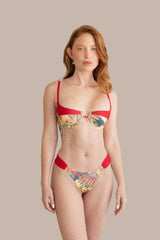 A woman modeling a two-piece bikini set with solid red straps and bottoms, and a multicolor tropical floral print on the cups and side ties, against a plain beige background.