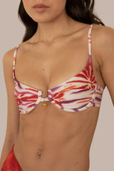 A close-up of a model wearing a bikini top with a white base and a vibrant red, purple, and orange abstract tie-dye pattern. The bikini top features a small gold ring detail at the center and thin shoulder straps.