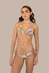 A woman modeling a two-piece string bikini with a colorful tropical leaf print, featuring a central gold-tone ring on the top and side-tie bottoms, set against a plain beige background.